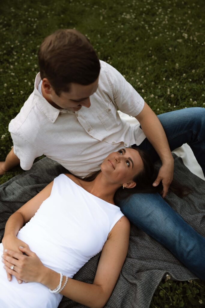 Woman lays on her fiance's lap in the middle of a lush green field.