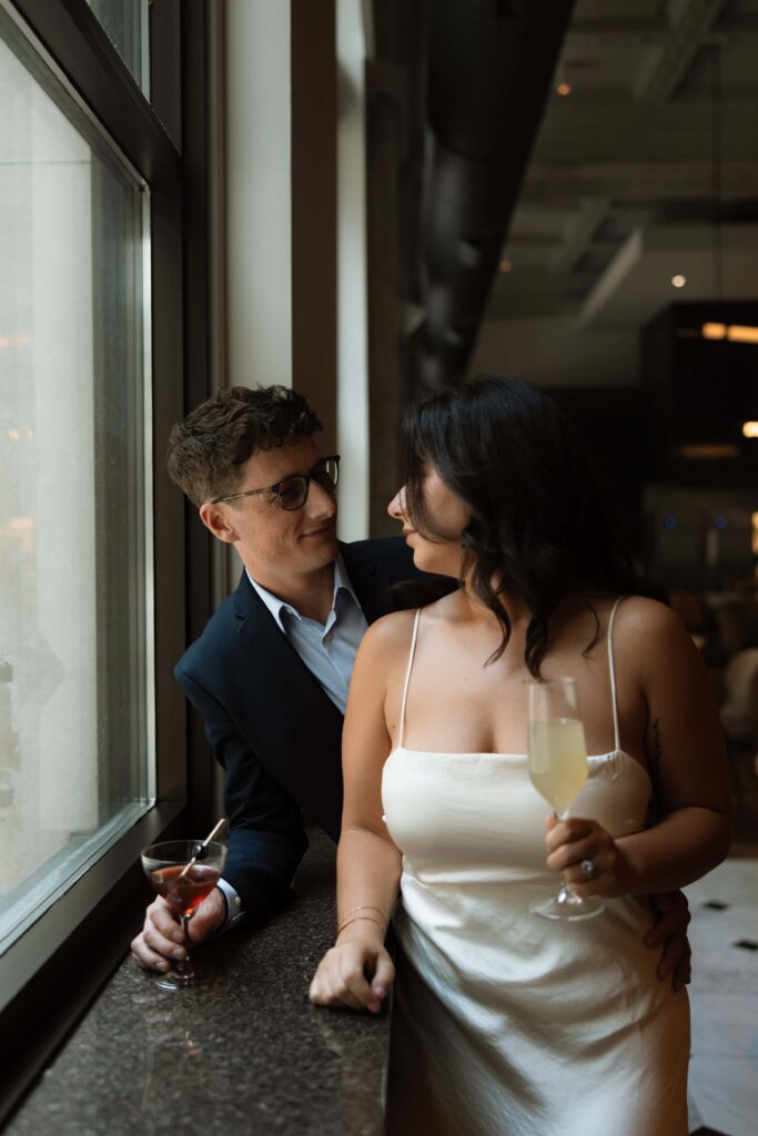 Engaged couple look at each other while holding their cocktails.