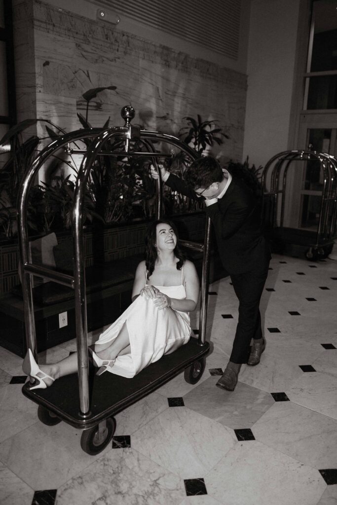 Woman sits on a luggage cart as her fiance pushes her at one of the best engagement session locations in the twin cities.