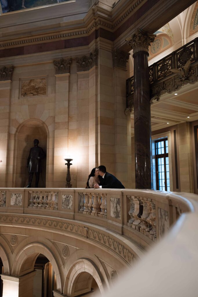 Romantic couple kiss as they lean on a marble railing at the St. Paul Capitol Building.