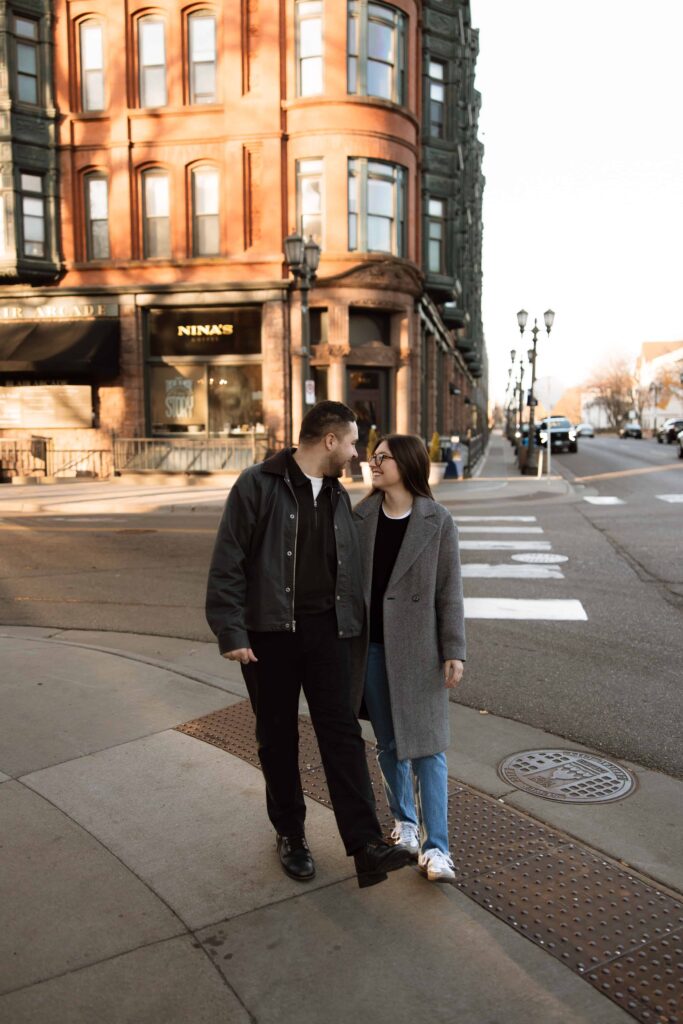Engaged couple walk in front of Nina's Cafe in St. Paul, Minnesota as they smile at each other.