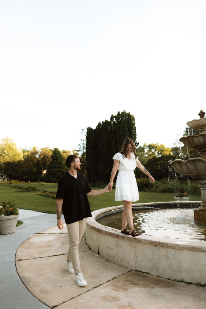 Woman walks on a fountain's ledge as her fiance helps her.
