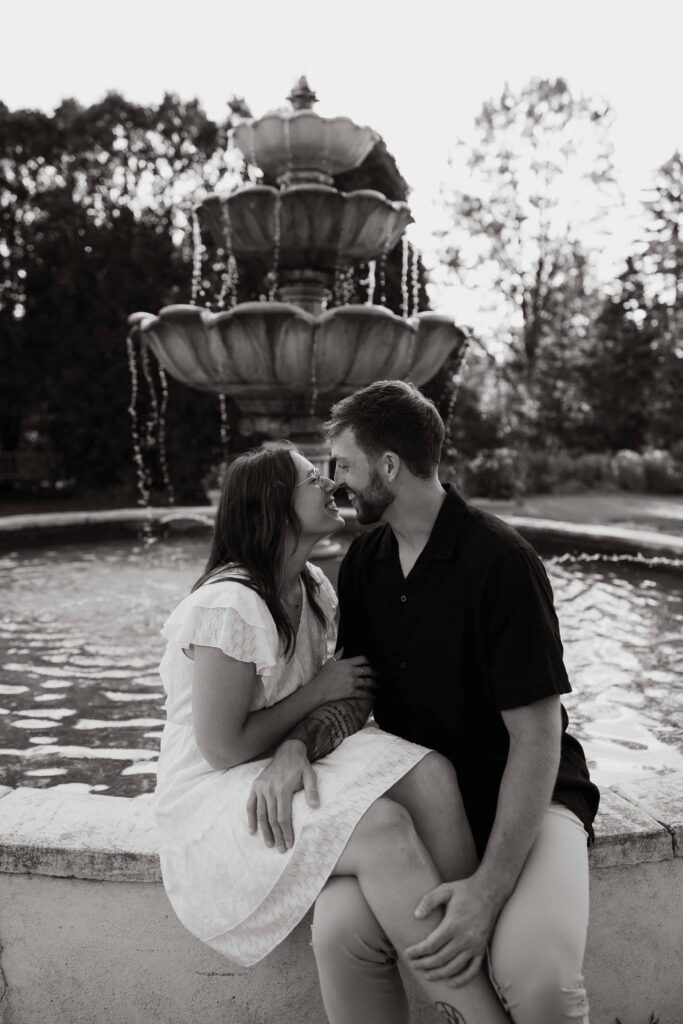 Romantic couple smiles at each other as they sit on the ledge of a fountain.