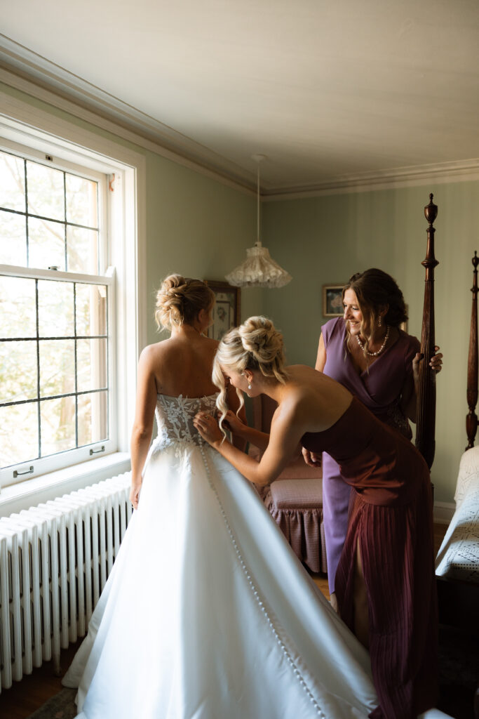 Sister of the bride and mother of the bride help the bride into her dress as she stands in front of a window.