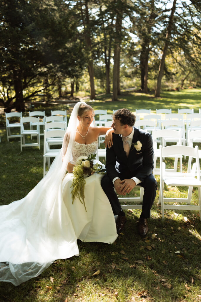 Bride and groom smile at each other as they sit on the ceremony chairs.