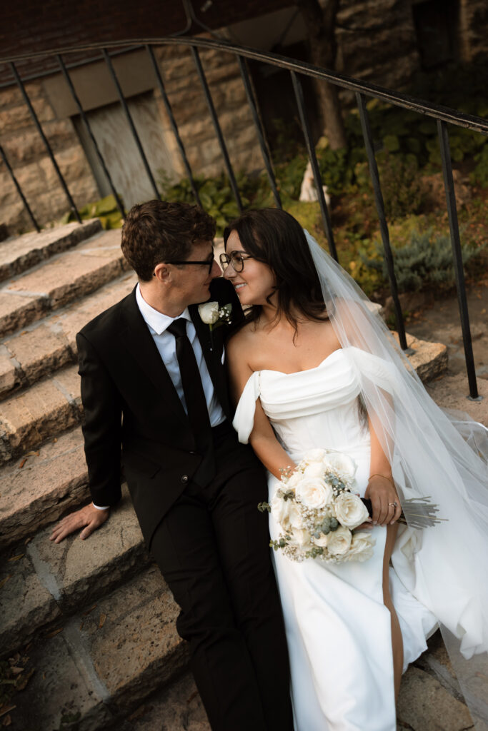 Bride and groom sit on steps as they smile at each other during their wedding portraits in Stillwater, Minnesota.