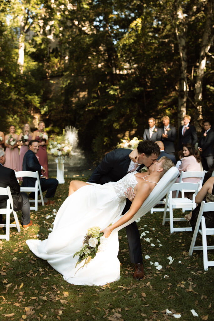 Bride and groom dip in the aisle after becoming husband and wife surrounded by lush greenery and flowers. 