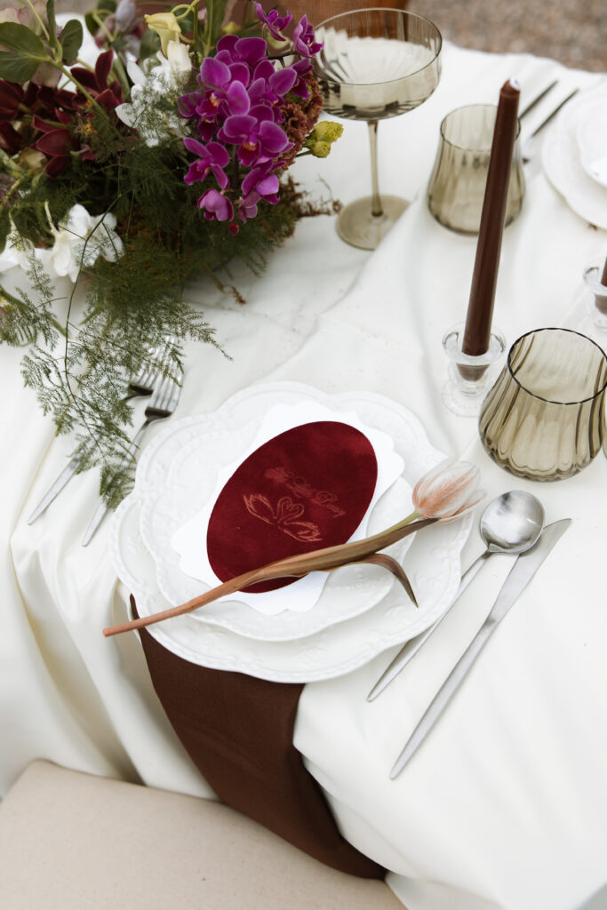 An intentional wedding reception place setting featuring a velvet menu card, single tulip stem, smoked glassware, and deep burgundy taper candles alongside lush orchid florals. An elevated, editorial wedding detail shot capturing the refined and romantic tablescape at The Little Daisy in Jerome, Arizona.