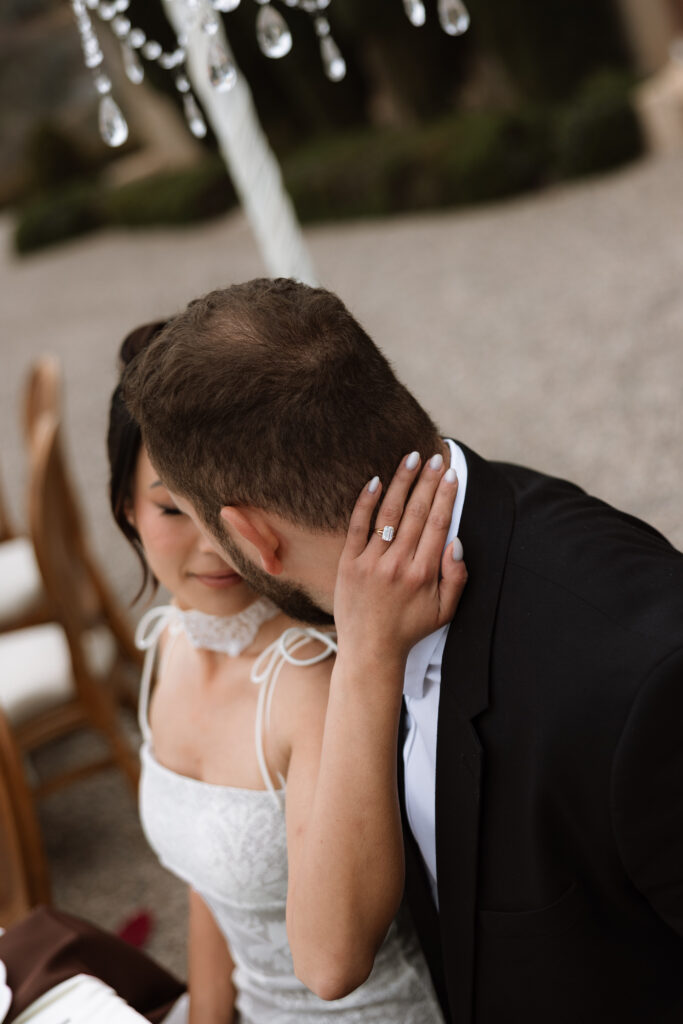 An intimate close-up of a newlywed couple seated together at The Little Daisy in Jerome, Arizona. The bride rests her hand on the groom’s neck, her engagement ring in focus, as they lean in close beneath softly blurred café lights and crystal chandeliers.