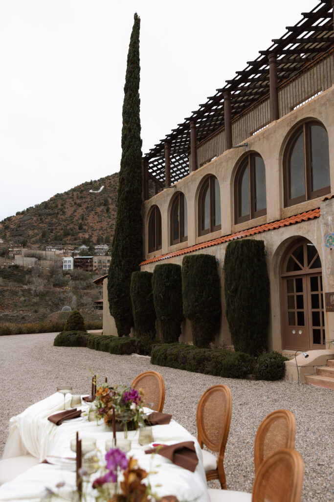 An outdoor wedding reception tablescape with linen linens, rattan chairs, and jewel-toned florals set against the Mediterranean-style arched facade of The Little Daisy in Jerome, Arizona. A romantic destination wedding reception with sweeping mountain views and timeless European-inspired architecture.