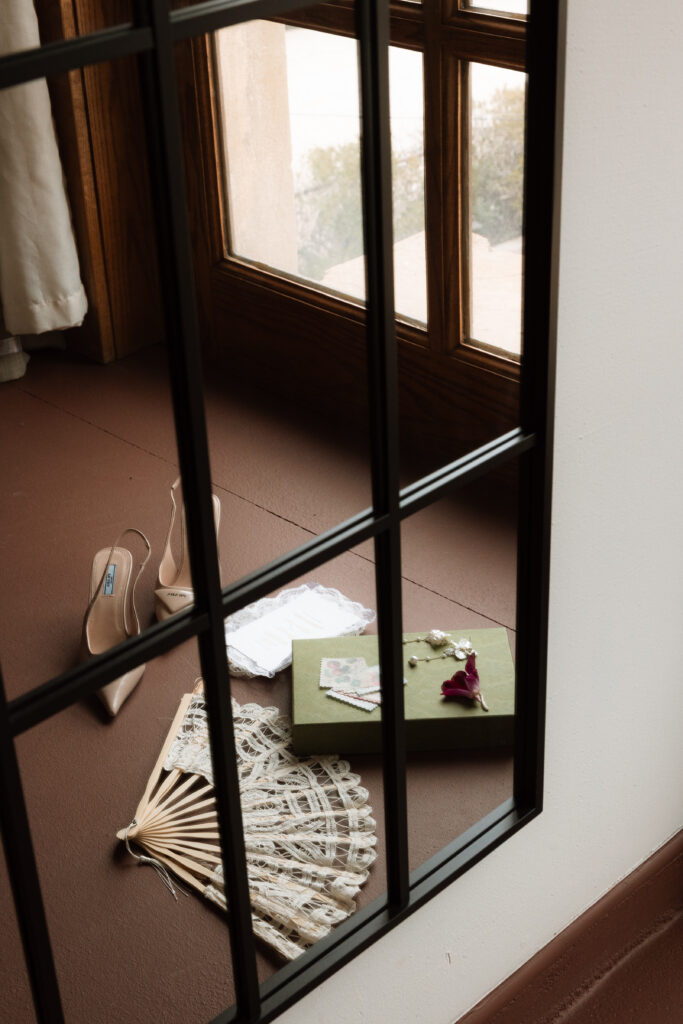 Bridal details including Prada heels, a lace fan, Gucci gift box, and delicate earrings reflected in a floor mirror with warm natural window light at The Little Daisy in Jerome, Arizona. An editorial wedding detail shot with timeless, intentional composition.