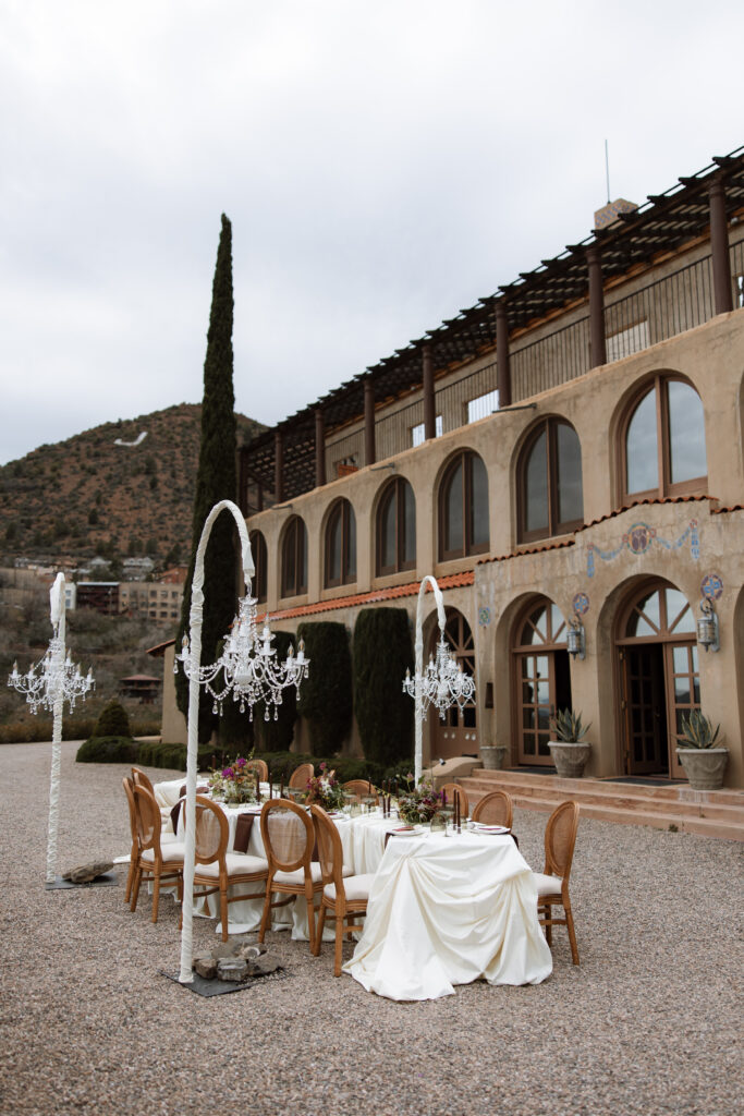 An outdoor wedding reception tablescape with freestanding crystal chandeliers, rattan chairs, and lush floral centerpieces set against the Mediterranean arched facade of The Little Daisy in Jerome, Arizona. A romantic, elevated destination wedding reception with sweeping mountain views.