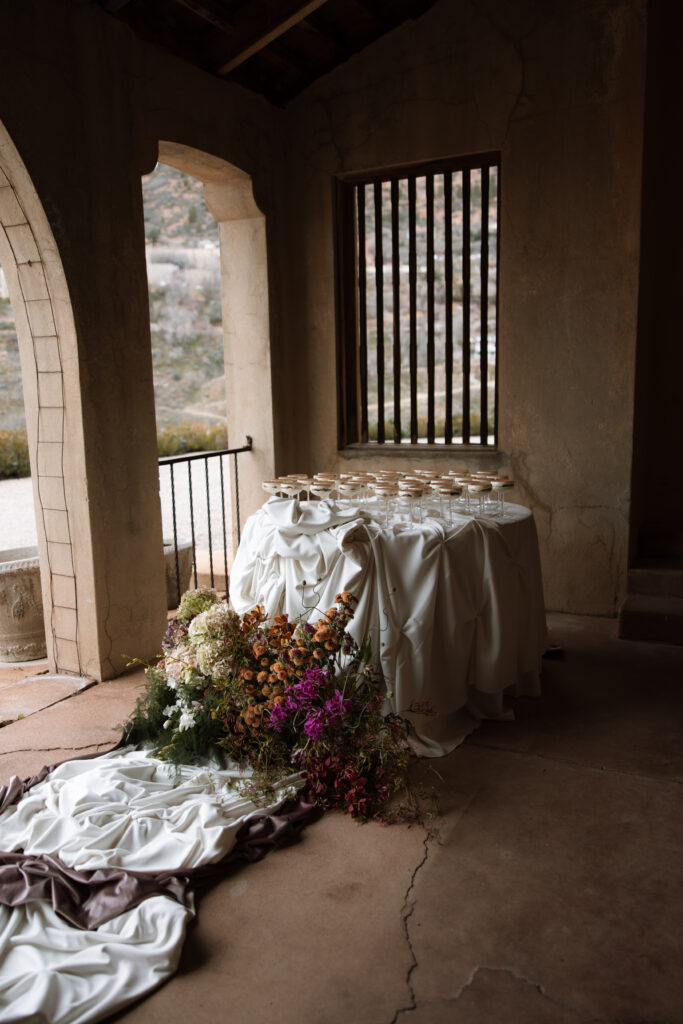 A draped champagne coupe display table adorned with a lush floor floral installation of jewel-toned and amber blooms beneath the stone arched loggia of The Little Daisy in Jerome, Arizona. An intimate, editorial wedding cocktail hour detail with moody, romantic atmosphere.