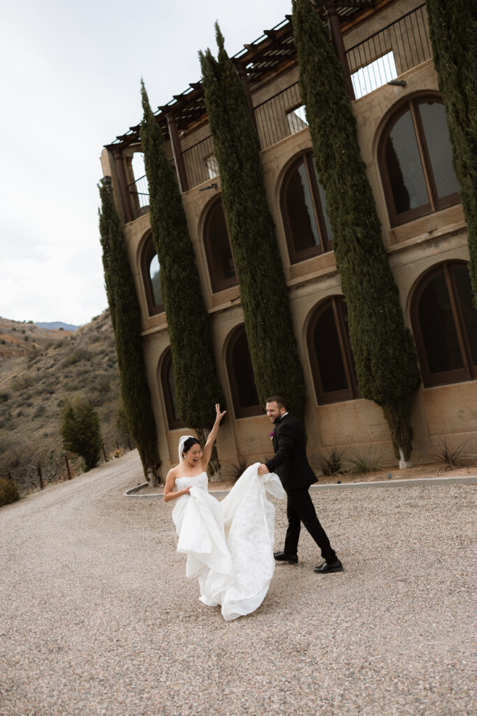 A joyful newlywed couple walks hand-in-hand across a gravel courtyard at The Little Daisy in Jerome, Arizona, the bride lifting her dress as she raises her arm in celebration. Tall cypress trees and arched windows line the historic hillside venue, with muted desert mountains and an overcast sky creating a soft, editorial backdrop.