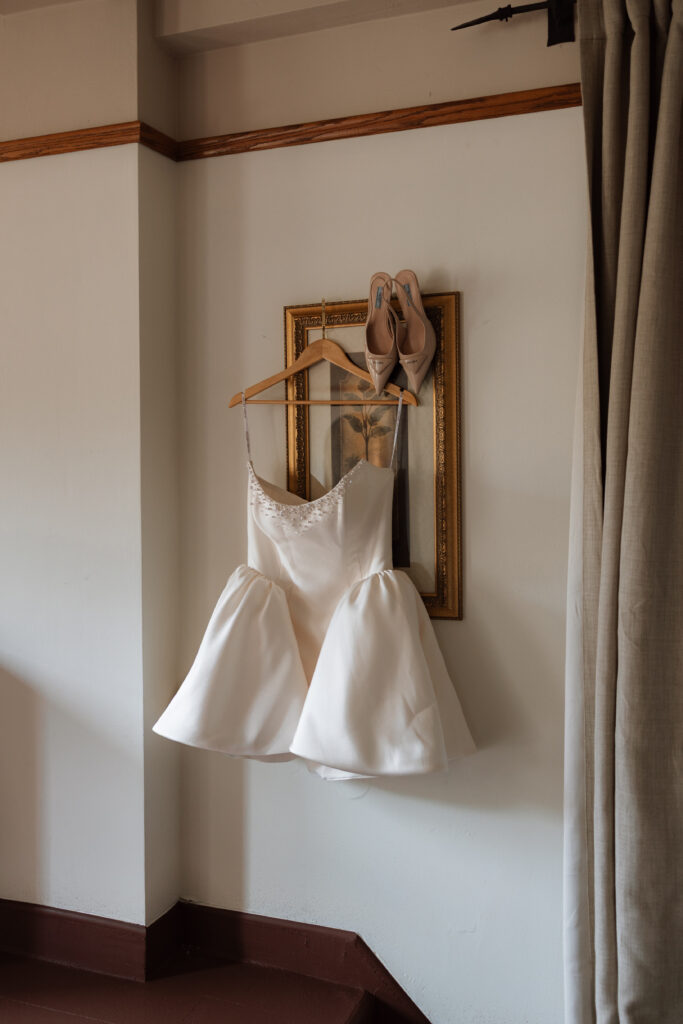 A short satin bridal reception dress with beaded detailing hanging on a wooden hanger against a gold-framed botanical print, with Prada heels resting on the frame. An editorial getting ready detail shot captured at The Little Daisy in Jerome, Arizona.