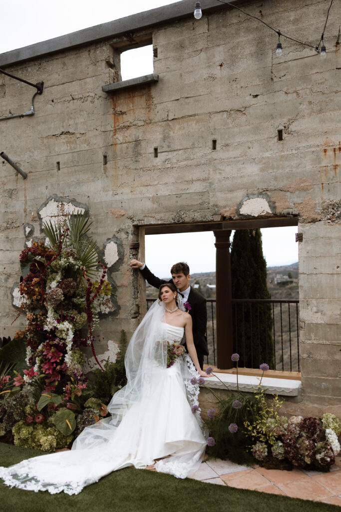 A bride and groom posing in the open-air ruins courtyard of The Little Daisy in Jerome, Arizona, surrounded by a lush, moody floral installation of burgundy, blush, and greenery with sweeping Arizona mountain views beyond. A dramatic, editorial destination wedding portrait with timeless elegance.