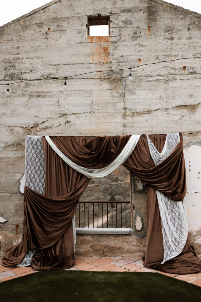 A draped wedding ceremony backdrop of rich mocha fabric and lace panels set against the historic concrete ruins of The Little Daisy in Jerome, Arizona. A moody, editorial ceremony altar with sweeping Arizona mountain views framed through the open wall.