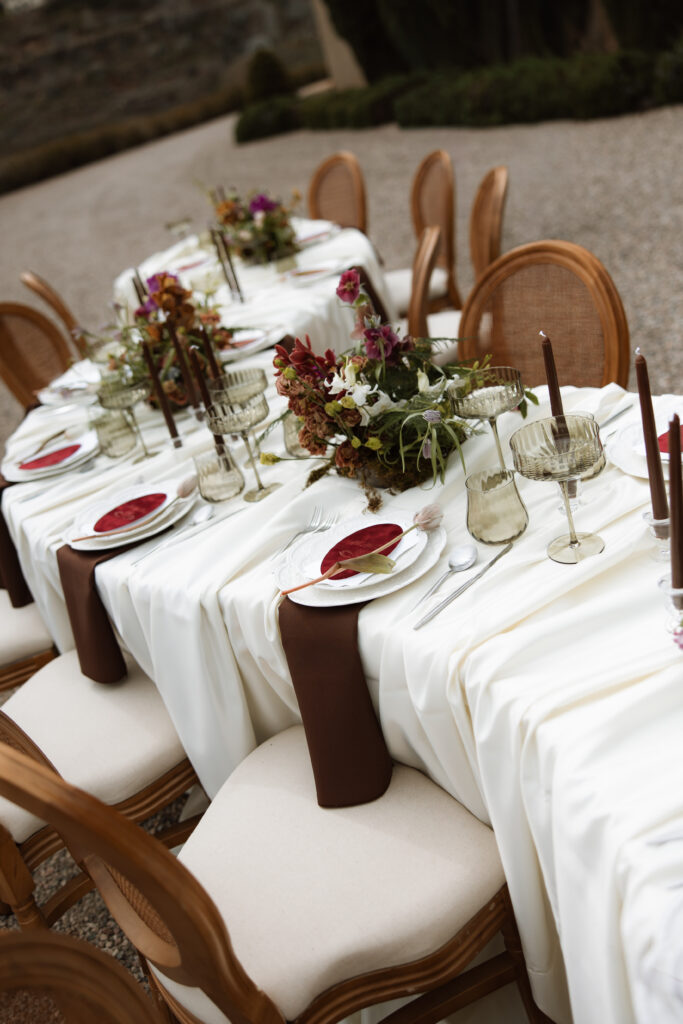 An elegantly styled outdoor reception table at The Little Daisy Jerome, Arizona, featuring warm wood cane-back chairs, deep burgundy tapered candles, smoked glass coupes, and lush floral centerpieces in rich jewel tones. A stunning detail shot from this historic Jerome wedding venue, perfect inspiration for couples planning an intentional destination wedding in the Southwest.